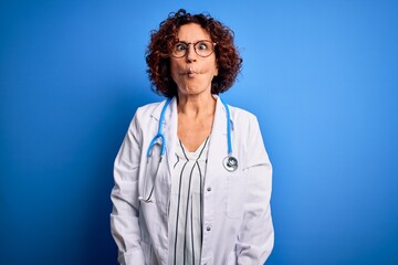 Sticker - Middle age curly hair doctor woman wearing coat and stethoscope over blue background making fish face with lips, crazy and comical gesture. Funny expression.