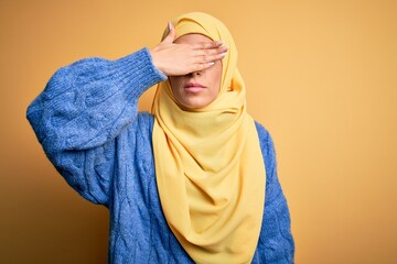 Canvas Print - Young beautiful brunette muslim woman wearing arab hijab over isolated yellow background covering eyes with hand, looking serious and sad. Sightless, hiding and rejection concept