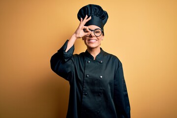 Canvas Print - Young beautiful brunette chef woman wearing cooker uniform and hat over yellow background doing ok gesture with hand smiling, eye looking through fingers with happy face.