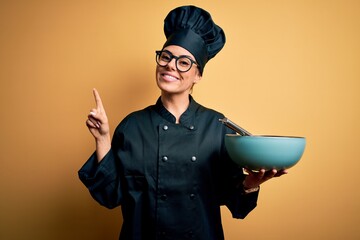 Wall Mural - Young beautiful brunette chef woman wearing cooker uniform and hat holding bowl and whisk surprised with an idea or question pointing finger with happy face, number one