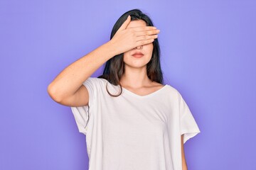 Poster - Young beautiful brunette woman wearing casual white t-shirt over purple background covering eyes with hand, looking serious and sad. Sightless, hiding and rejection concept