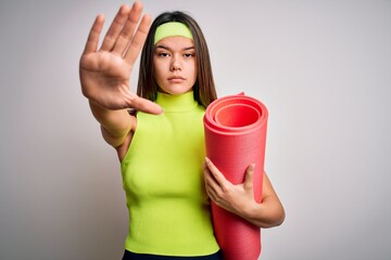 Wall Mural - Beautiful sporty girl doing sport wearing sportswear holding yoga mat over white background with open hand doing stop sign with serious and confident expression, defense gesture