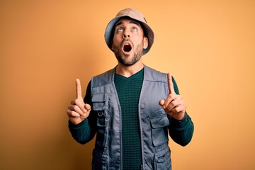 Canvas Print - Handsome tourist man with beard on vacation wearing explorer hat over yellow background amazed and surprised looking up and pointing with fingers and raised arms.