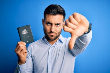 Canvas Print - Young handsome tourist man holding australia australian passport id over blue background with angry face, negative sign showing dislike with thumbs down, rejection concept