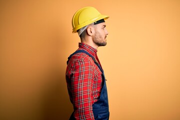 Canvas Print - Young builder man wearing construction uniform and safety helmet over yellow isolated background looking to side, relax profile pose with natural face with confident smile.