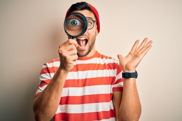 Young detective man looking through magnifying glass over isolated background very happy and excited, winner expression celebrating victory screaming with big smile and raised hands