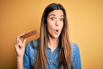 Young beautiful girl holding healthy protein bar standing over isolated yellow background scared in shock with a surprise face, afraid and excited with fear expression