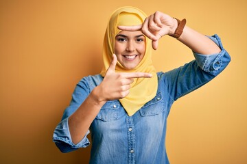 Poster - Young beautiful girl wearing muslim hijab standing over isolated yellow background smiling making frame with hands and fingers with happy face. Creativity and photography concept.