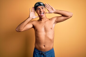 Poster - Young handsome man shirtless wearing swimsuit and swim cap over isolated yellow background Smiling cheerful playing peek a boo with hands showing face. Surprised and exited