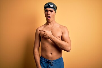 Poster - Young handsome man shirtless wearing swimsuit and swim cap over isolated yellow background Surprised pointing with finger to the side, open mouth amazed expression.