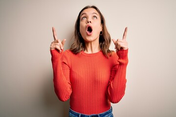 Young blonde girl wearing casual red sweater over isolated background amazed and surprised looking up and pointing with fingers and raised arms.