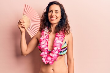 Young beautiful hispanic woman wearing bikini and hawaiian lei holding hand fan looking positive and happy standing and smiling with a confident smile showing teeth