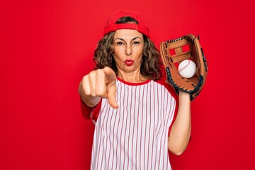 Middle age senior woman wearing baseball equiment, ball and glove over red isolated background pointing with finger to the camera and to you, hand sign, positive and confident gesture from the front