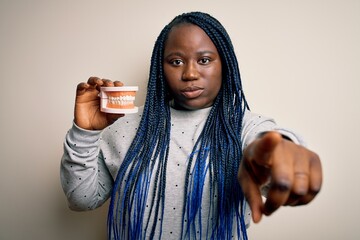 Sticker - African american plus size woman with braids holding plastic teeth over white background pointing with finger to the camera and to you, hand sign, positive and confident gesture from the front