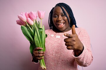 Sticker - Young african american plus size woman with braids holding bouquet of pink tulips flower pointing fingers to camera with happy and funny face. Good energy and vibes.