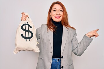 Wall Mural - Young beautiful redhead woman holding bag with money and dollar sign over white background very happy pointing with hand and finger to the side