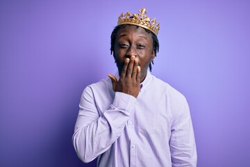 Poster - Young african american man wearing golden crown of king over isolated purple background bored yawning tired covering mouth with hand. Restless and sleepiness.