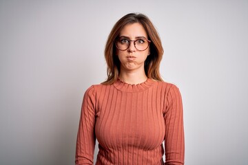 Young beautiful brunette woman wearing casual sweater and glasses over white background puffing cheeks with funny face. Mouth inflated with air, crazy expression.