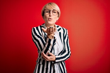 Poster - Blonde business woman with short hair wearing glasses and striped jacket over red background looking at the camera blowing a kiss with hand on air being lovely and sexy. Love expression.