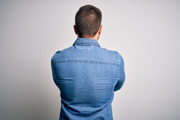 Young handsome blond man with beard and blue eyes wearing casual denim shirt standing backwards looking away with crossed arms