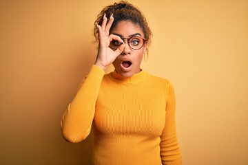 Sticker - Young beautiful african american girl wearing sweater and glasses over yellow background doing ok gesture shocked with surprised face, eye looking through fingers. Unbelieving expression.