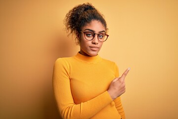 Sticker - Young beautiful african american girl wearing sweater and glasses over yellow background Pointing with hand finger to the side showing advertisement, serious and calm face