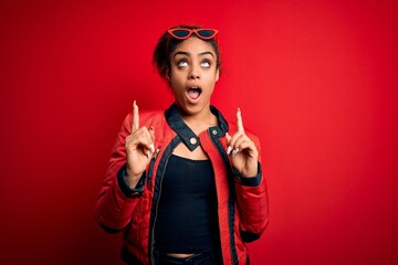 Poster - Beautiful african american girl wearing red jacket and sunglasses over isolated background amazed and surprised looking up and pointing with fingers and raised arms.