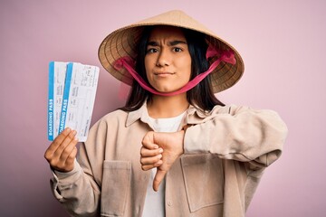 Canvas Print - Young tourist woman on vacation wearing traditional asian hat holding airplane boarding pass with angry face, negative sign showing dislike with thumbs down, rejection concept