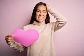 Canvas Print - Young beautiful asian woman holding pink heart standing over isolated background stressed with hand on head, shocked with shame and surprise face, angry and frustrated. Fear and upset for mistake.