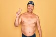 © Krakenimages.com - Middle age senior grey-haired swimmer man wearing swimsuit, cap and goggles smiling looking to the camera showing fingers doing victory sign. Number two.