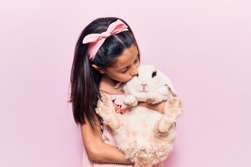 Adorable hispanic kid girl wearing diadem smiling happy. Holding and kissing cute rabbit standing over isolated pink background