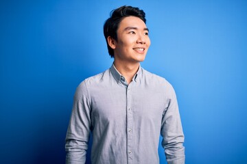 Wall Mural - Young handsome chinese man wearing casual shirt standing over isolated blue background looking away to side with smile on face, natural expression. Laughing confident.