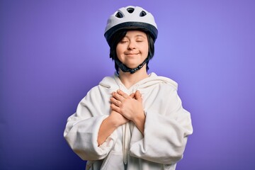 Sticker - Young down syndrome cyclist woman wearing security bike helmet over purple background smiling with hands on chest with closed eyes and grateful gesture on face. Health concept.