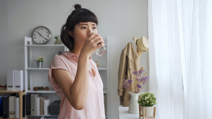 attractive young asian japanese woman drinking fresh water at home living room. beautiful girl holding glass of fresh healthy beverage and enjoy sunshine through window in cozy apartment living room