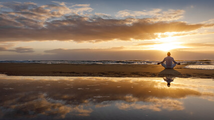 yoga meditation silhouette lotus sunrise beach, mindfulness, wellness and wellbeing concept, water reflection of man in yoga lotus pose sitting alone on sand with ocean cloud background, copy space