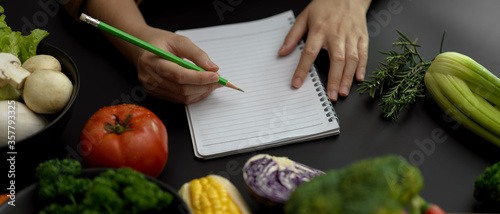 Fotomural Female student taking recipe notes on blank notebook on black kitchen table in c