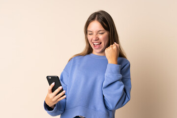 Young Lithuanian woman using mobile phone isolated on beige background celebrating a victory