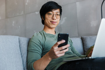 Poster - Image of pleased asian man using cellphone and laptop while sitting