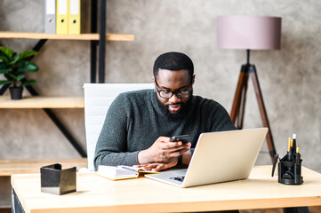 An african-american guy in smart casual wear and glasses is typing messages on smartphone while working with a laptop in contemporary office