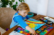 © Irina Schmidt - little toddler girl painting rainbow and sun with water colors during pandemic coronavirus quarantine disease. Children painting rainbows around the world with the words Let's all be well. Happy child