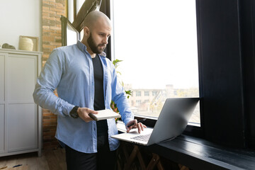 A guy in a blue shirt is standing by the window working on a laptop