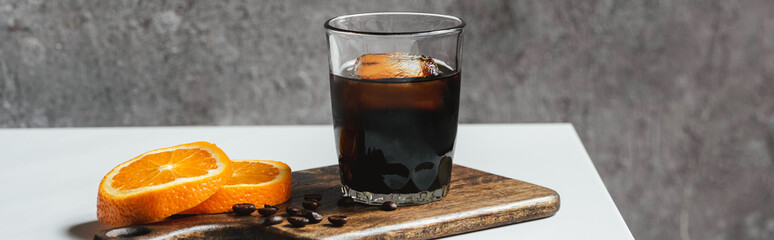 Wall Mural - cold brew coffee with ice in glass near orange slices on chopping board and coffee beans on white table, panoramic shot