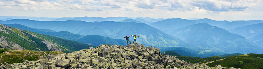 Panoramic view of loving couple holding hands up beautiful mountains scenery on background. Mountain hiking, tourists reaching peak together. Wild nature with amazing views. Sport tourism concept.