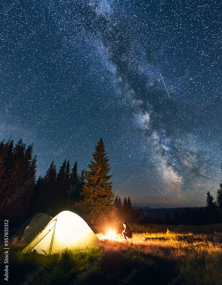 Lonely guy sitting near campfire in campsite under starry sky in the mountains. Night sky is strewn with bright stars and prominent Milky way. Tourist tents in valley with large pine trees. - obrazy, fototapety, plakaty 