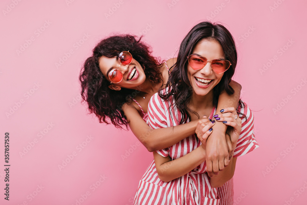 Young, spectacular models vigorously pose for portrait in studio. Tanned sisters embrace and laugh in unusual glasses - obrazy, fototapety, plakaty 