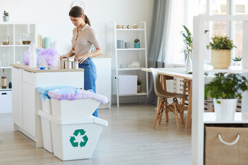 Young woman standing at the kitchen table and recycling plastic bottles and can