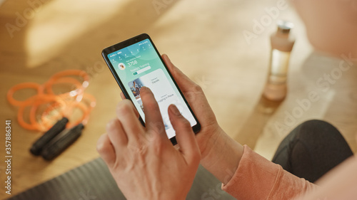 Fotomural Over Shoulder Shot: Fit Woman Holds Smartphone, Uses Fitness Tracking App, Uses Gesture to Choose Premium Workout Videos