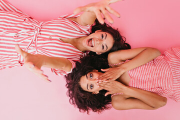 Girlfriends with dark curly hair are having fun at their pajama party. Laughing girl stretches towards camera while her cousin covers her face in surprise