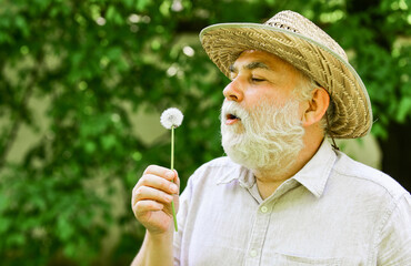 Peacefulness. Tranquility and serenity. Harmony of soul. Elderly man in straw summer hat. Happy and carefree retirement. Lonely grandpa blowing dandelion seeds in park. Mental health. Peace of mind