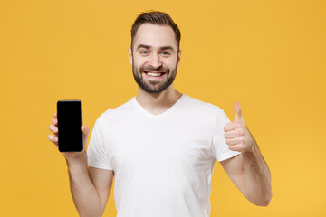Smiling young bearded man guy in white casual t-shirt isolated on yellow background studio. People lifestyle concept. Mock up copy space. Hold mobile phone with blank empty screen, showing thumb up.